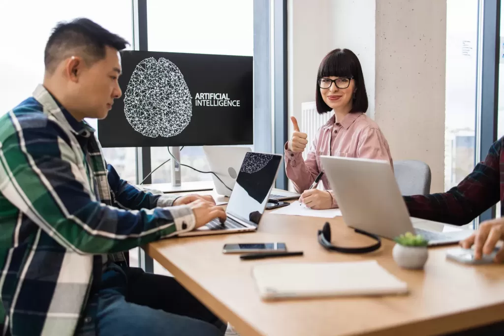 Three people work at a table with laptops; a screen displays an artificial intelligence graphic, and one woman gives a thumbs up.