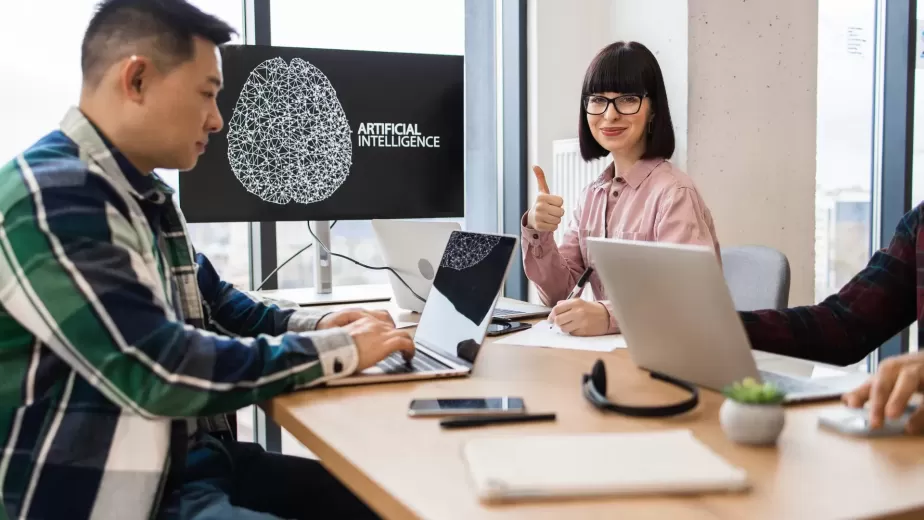 Three people work at a table with laptops; a screen displays an artificial intelligence graphic, and one woman gives a thumbs up.