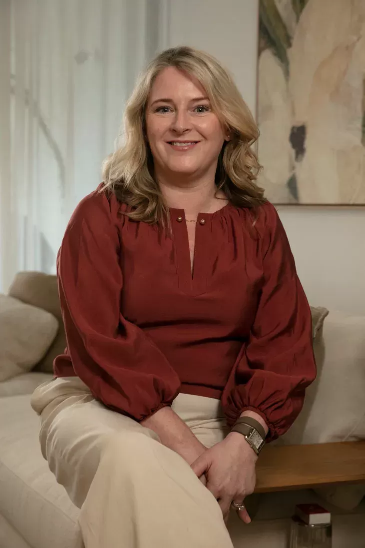 A woman with blonde hair wearing a maroon blouse and light pants sits on a beige couch, smiling at the camera in a softly lit living room.