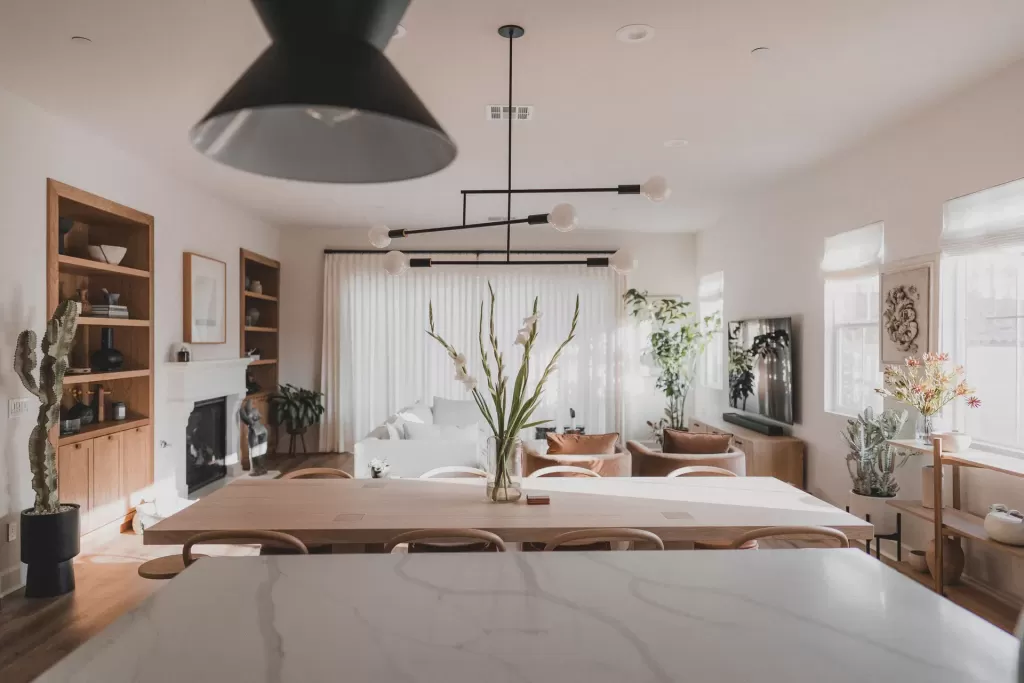 A modern, well-lit living and dining area with a marble kitchen island, wooden furniture, neutral tones, large windows, plants, and contemporary light fixtures.