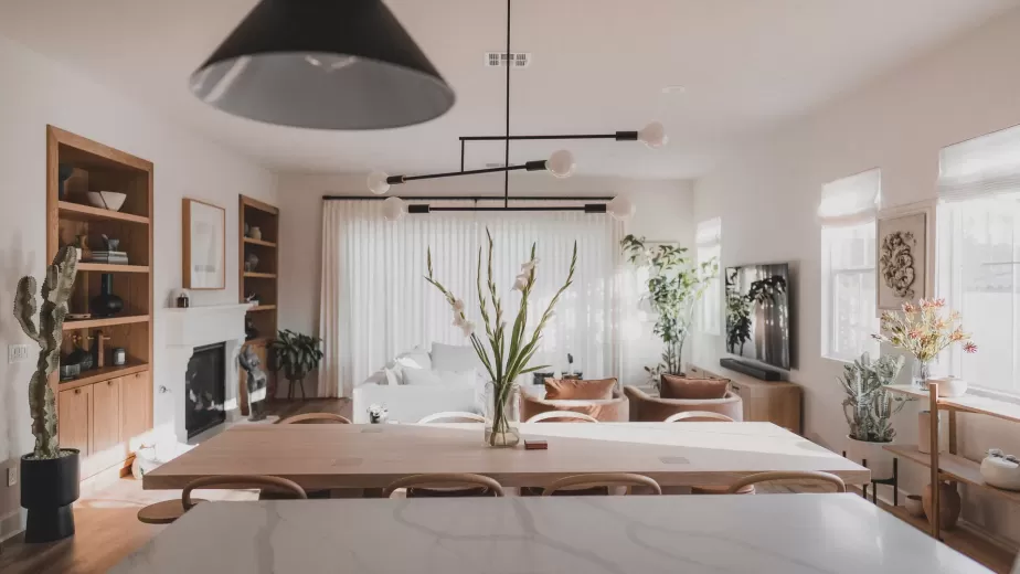 A modern, well-lit living and dining area with a marble kitchen island, wooden furniture, neutral tones, large windows, plants, and contemporary light fixtures.
