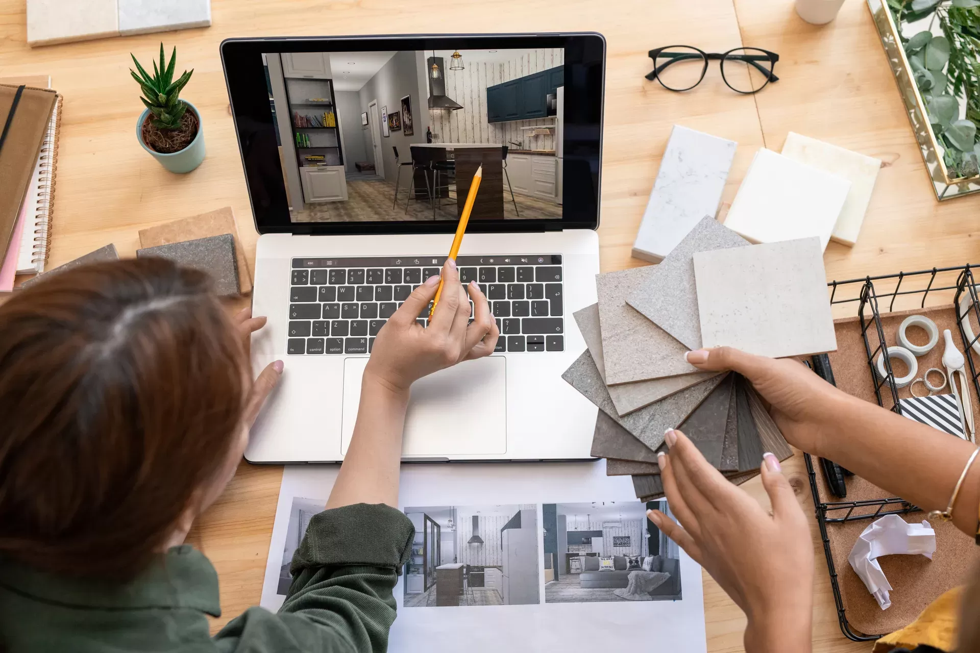 Two people review flooring samples and interior design plans, referencing a kitchen image on a laptop and printed layouts on a desk.