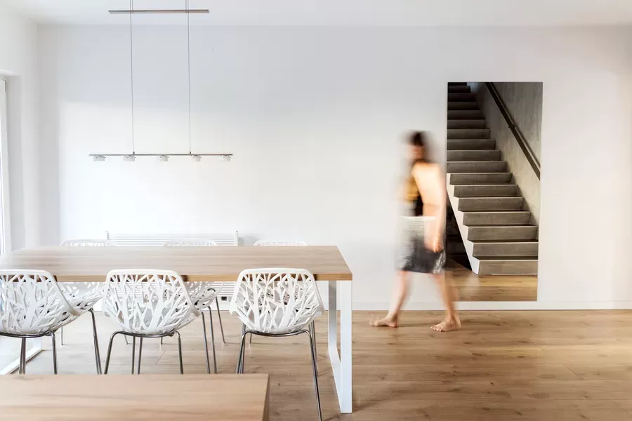 A person walks barefoot through a minimalist dining room with white chairs, a wooden table, and a staircase visible through an opening in the wall.