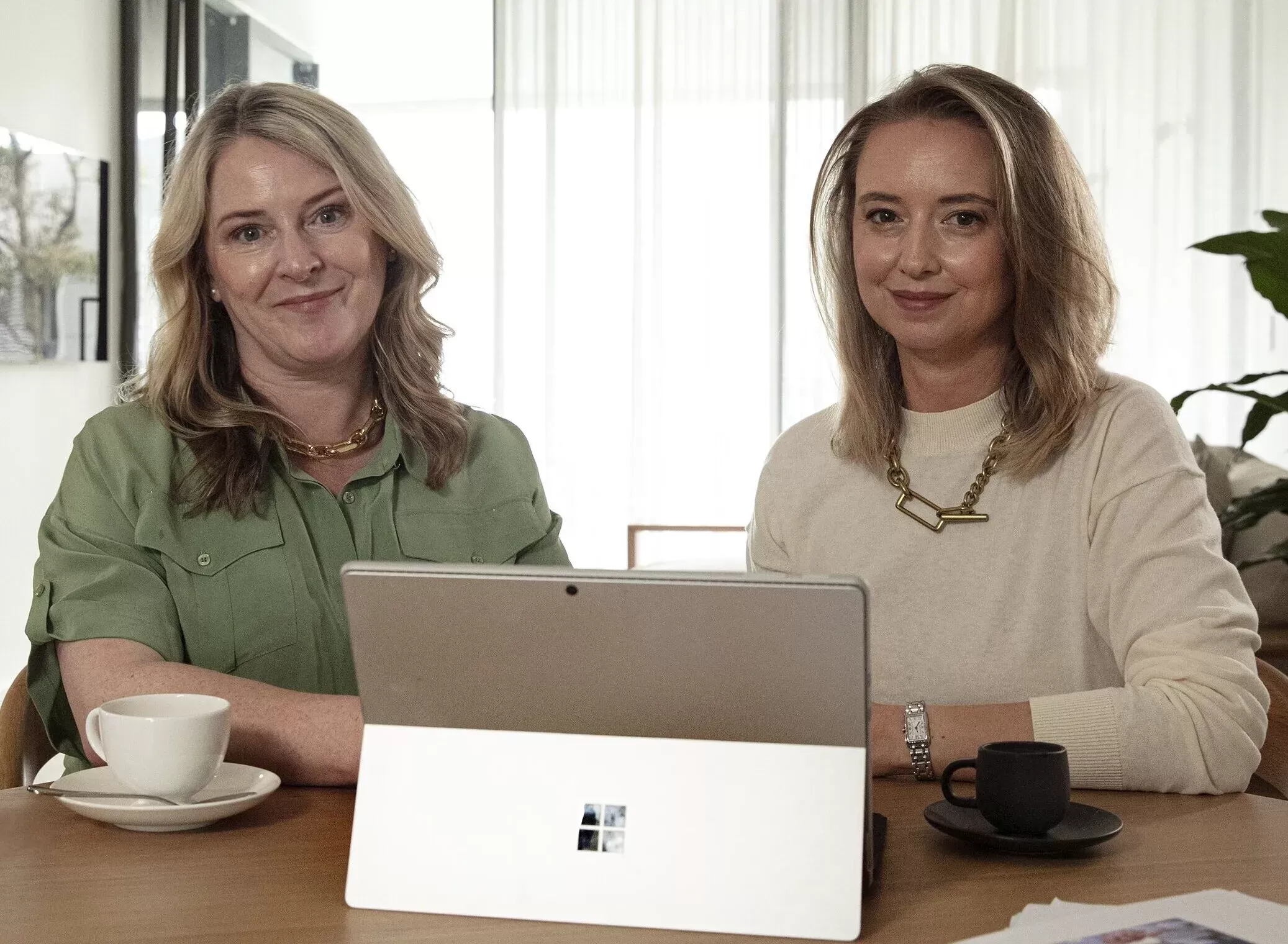Two women sit at a table with a Microsoft Surface laptop, each with a cup of coffee, facing the camera in a well-lit room.