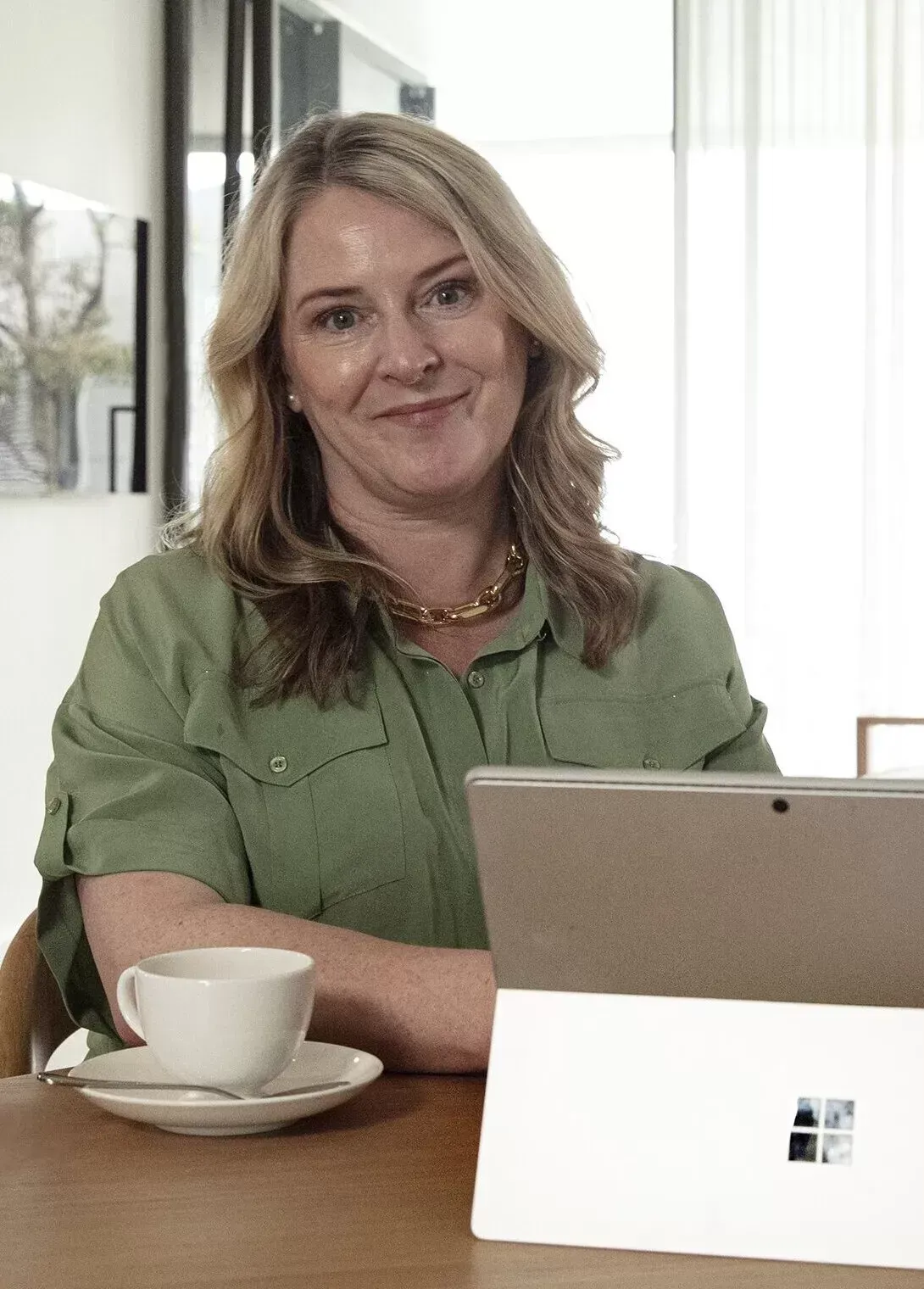 Two women sit at a table with a laptop and coffee cups, facing the camera in a well-lit, modern indoor setting.